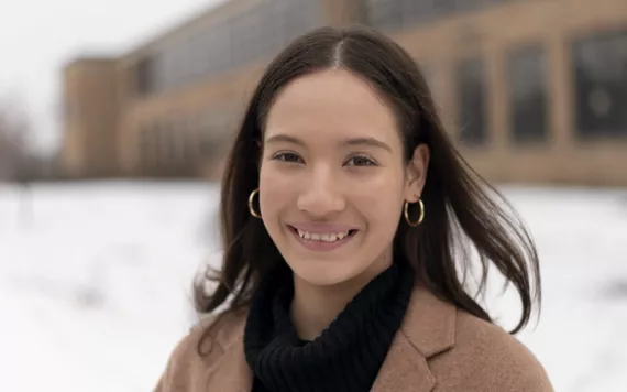 Alexandra Collins is smiling at the camera, wearing a brown coat and black turtleneck and standing in front of her high school with snow on the ground.