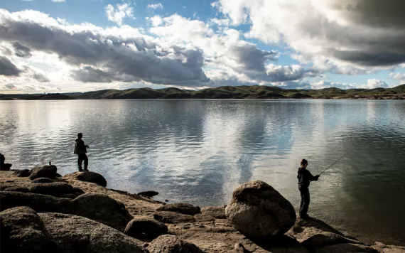 Two people are fishing from the shore of Millerton Lake. The water is glassy, and blue sky pokes through clouds above.