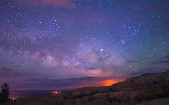 Image of an indigo pinkish sky with a setting or rising sun in the background, and visible stars and galaxies. 