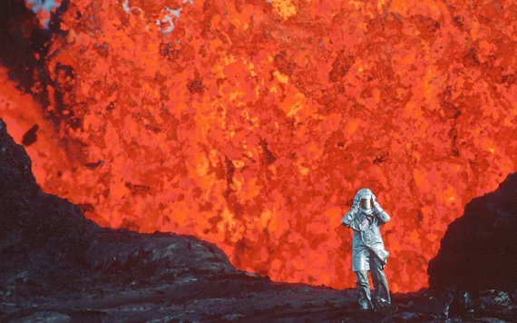 A small human figure in a silver foil jumpsuit and helmet stands in front of a fountain of hot red lava.