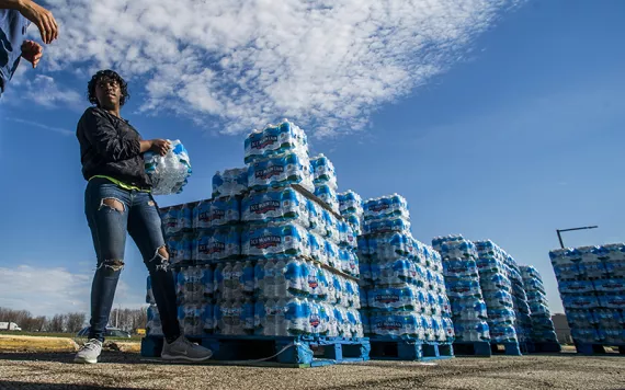 A woman in ripped jeans and a windbreaker hands a package of water to a person not in the photo. A pallet of bottled water is behind her.