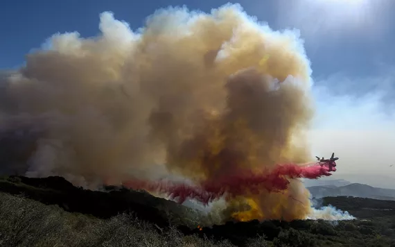 Cloud of fire retardent bursting over a green-brown hilly landscape that is on fire, against a blue sky.