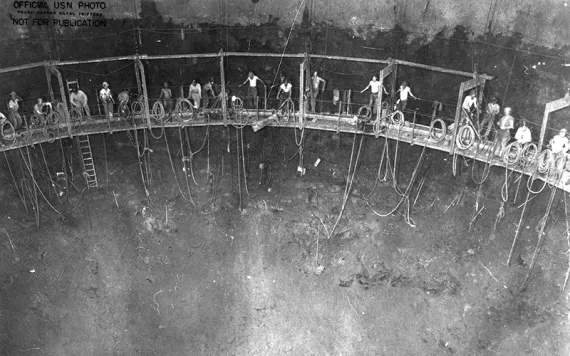 Black and white photo of tiny people in workwear on a narrow walkway inside a massive metal cavern of some kind.