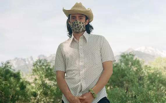 Andy Butler faces the camera with trees behind him. Butler is wearing a leopard-print face mask, a straw cowboy hat, and a light-colored button-up shirt.