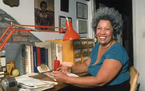 Portrait of American author Toni Morrison smiling and sitting at her desk at home, 1980s.