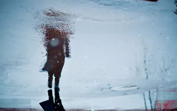 Blurry silhouette of a walking figure in a long coat, reflected onto a blue, rainy street.