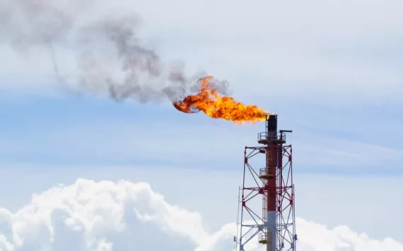 A bright orange flame and smoke coming out of an oil well against a light blue sky