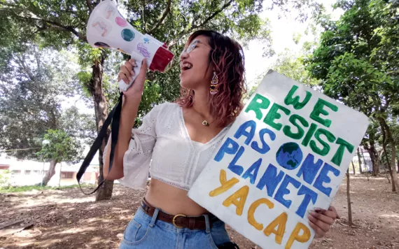 Mitzi Jonelle Tan is outside and talking into a megaphone. She's holding up a sign that says "We Resist As One Planet/YACAP"