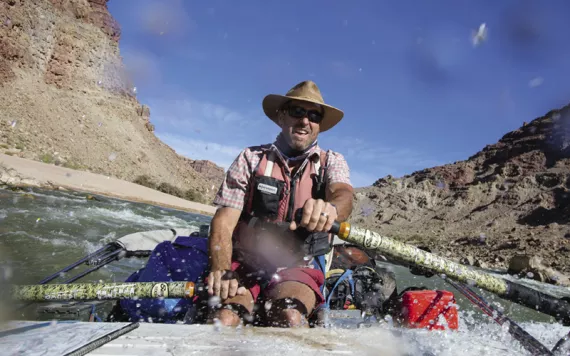 A man steers a boat on a river with two oars. Behind him is blue sky and desert/rock walls.