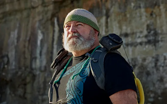 Wayne Thompson, who has a gray beard and is wearing a beanie and a backpack, looks off in the distance.