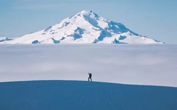 A hiker walks across a snowy ridge with a layer of fog and Glacier Peak in the distance.