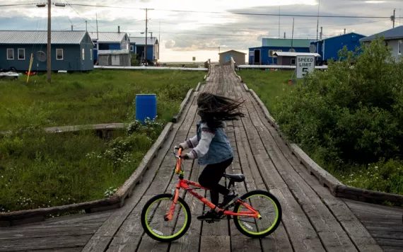 A kid with long hair covering their face rides a bike on a wooden boardwalk. A sign says "Speed Limit 5."