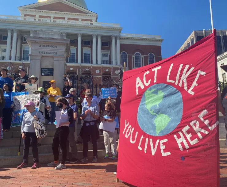 Rally in front of the MA State House with a large red banner that reads "Act like you live here" with a painting of the earth in the center.