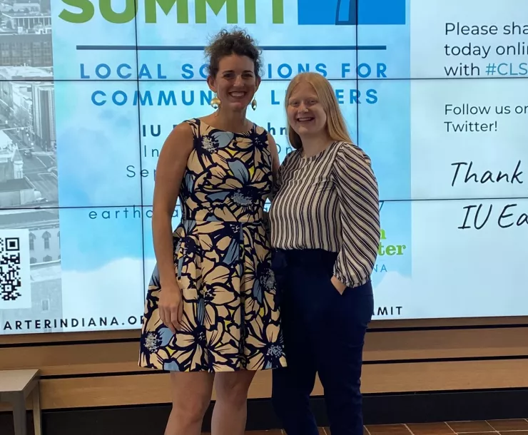 Two women stand in front of a large screen which says Climate Leadership Summit 7 on it. They are smiling and looking at the camera. The woman on the left has brown curly hair tied up and a floral dress. The woman on the right has long red hair, a stripy top, and dark pants.