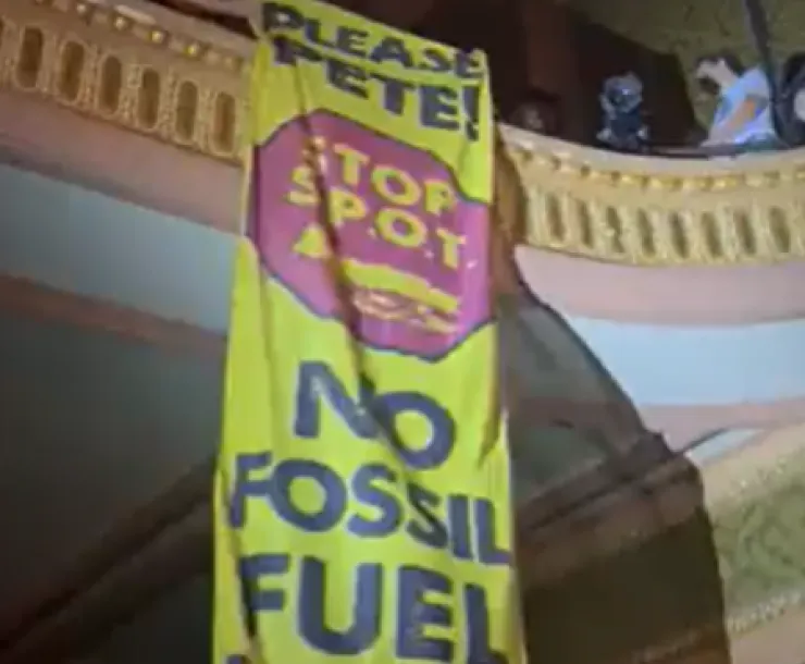 A long yellow banner reading "Please Pete! STOP S.P.O.T. No Fossil Fuel Exports!" is unfurled from a balcony.