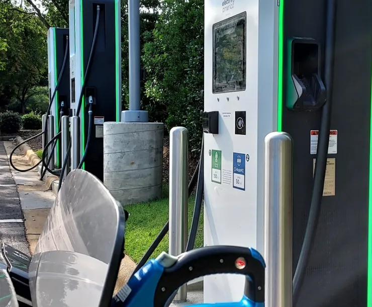 The hood of an electric car is shown with its charging port open and plugged into a charger in a parking lot surrounded by trees.