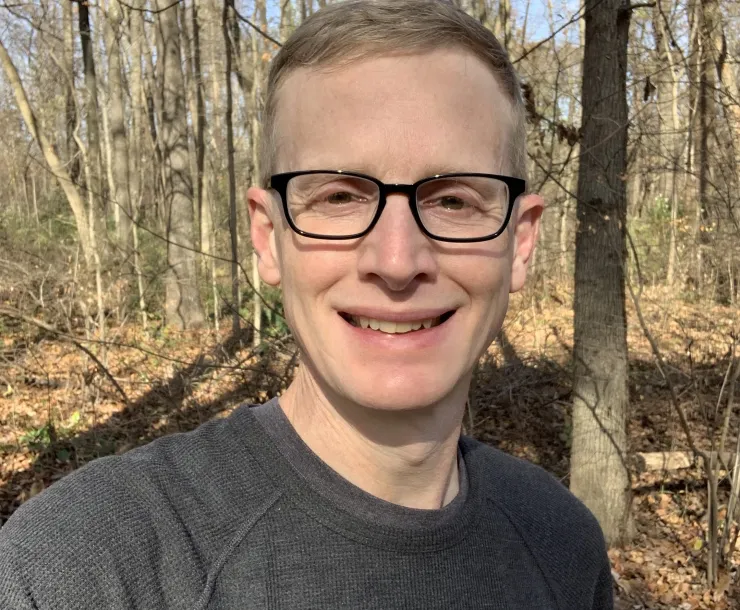 A man with short light brown hair and dark glasses stands with bare trees visibly behind him. He is smiling.