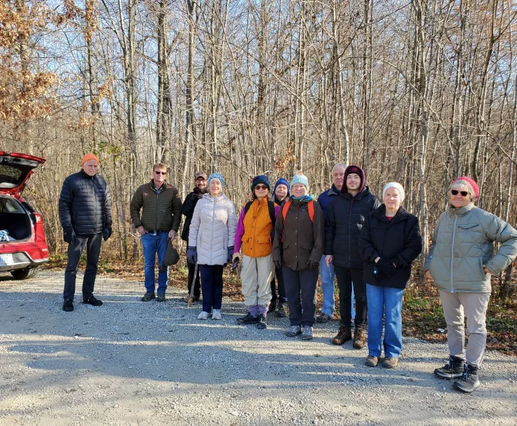 A group of people stand in a parking lot at a trail head of a forest, about to go on a hike. They are dressed for the cold weather and smiling.