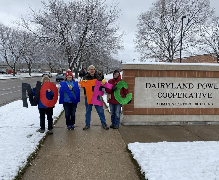 Four Coulee Region Group members stand in front of Dairyland Power Cooperative's headquarters in La Crosse, holding up colored letters that say "NO NTEC".