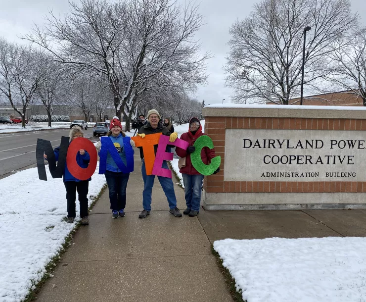 Four Coulee Region Group members stand in front of Dairyland Power Cooperative's headquarters in La Crosse, holding up colored letters that say "NO NTEC".
