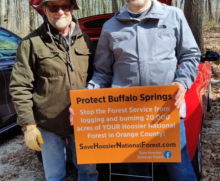 Two men stand in a parking lot with trees in the background. They are holding an orange sign which says Protect Buffalo Springs. Stop the Forest Service from logging and burning 20,000 acres of YOUR Hoosier National Forest in Orange County! SaveHoosierNationalForest.com