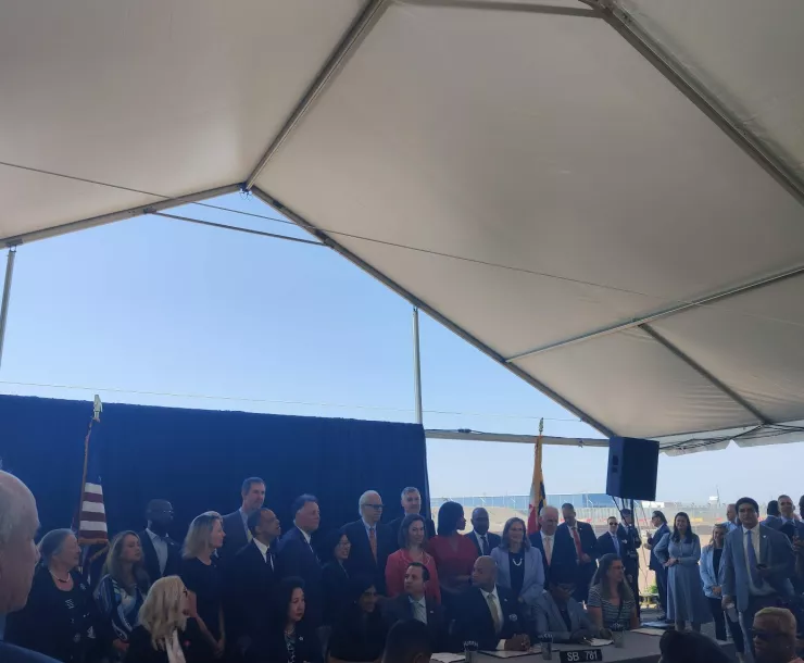 Dozens of legislators and agency staff gather around Governor under a canopy on a clear blue day