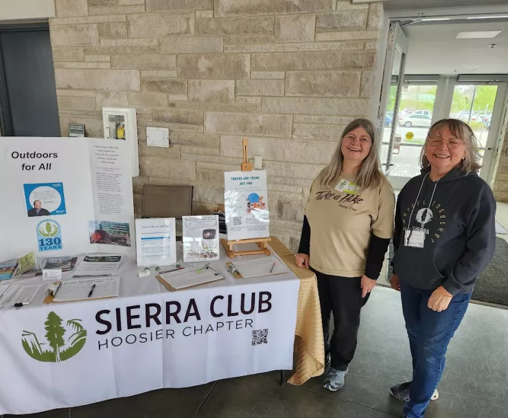 Two women standing next to a table with a white tablecloth with the Sierra Club Hoosier Chapter logo in green and black. There are various Sierra Club materials on the table, including a display about the Outdoors for All campaign.