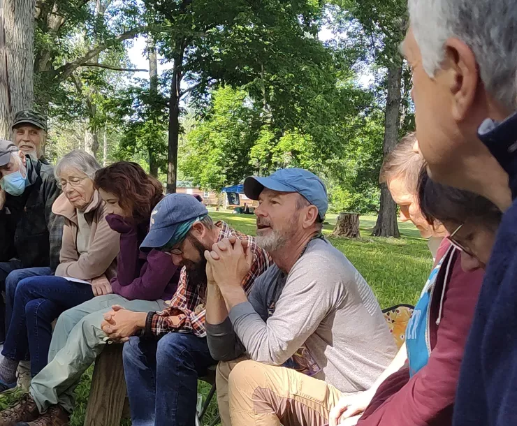 Nine people sitting outside. They appear to be forming part of a circle. The grass is green and so are the leaves on the trees in the background. A person with a blue baseball cap is speaking and has the attention of the other people in the circle.