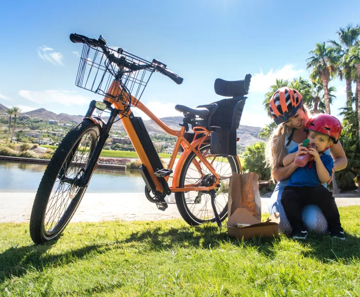 Mother and son wearing helmets and sitting next to an e-bike on some sunny grass.