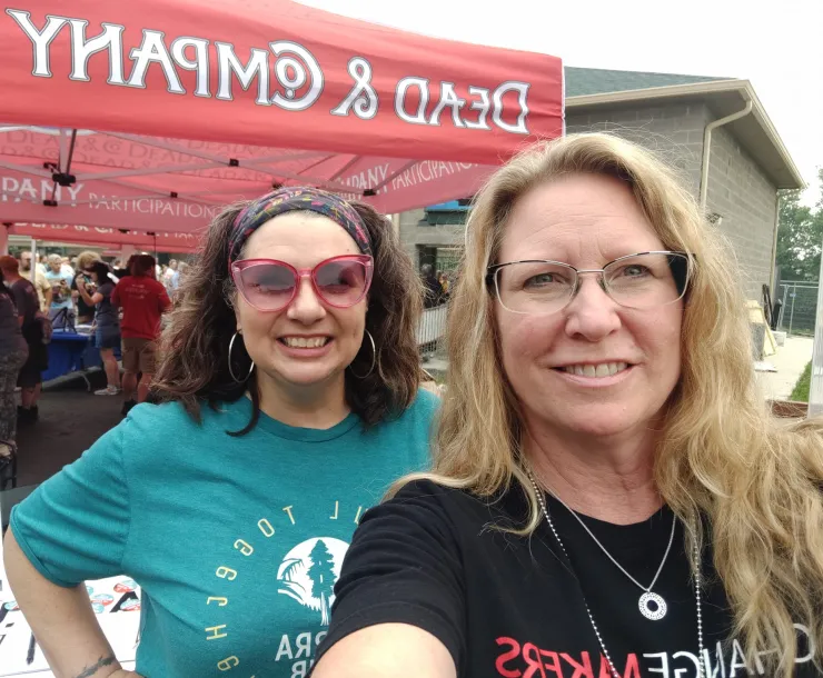 A selfie of two women standing outside at an event. One has blonde hair and the other brown hair. They are both smiling. There is a red canopy set up in the background with the words Dead & Company in white.