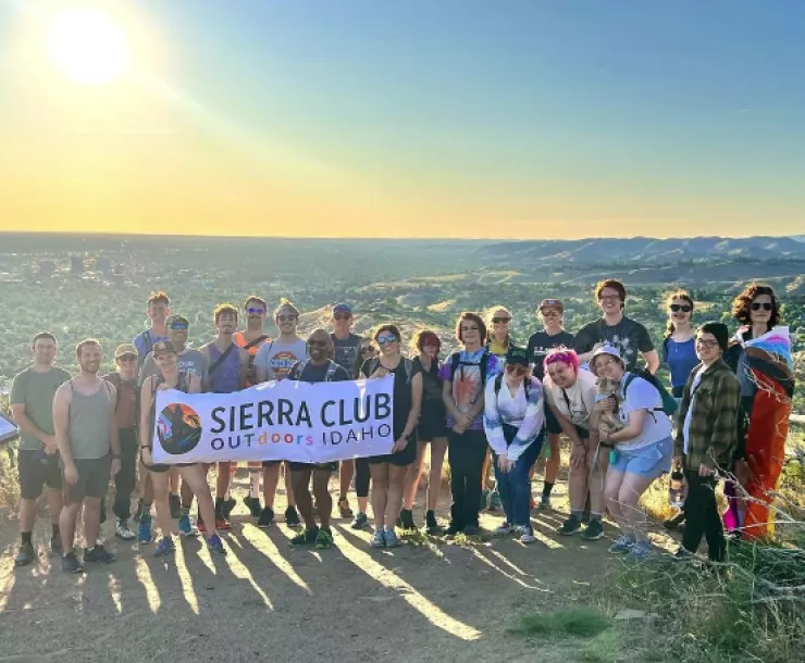 Picture of lots of people standing at top of a mountain overlooking a city holding a banner that says "Sierra Club OUTdoors Idaho" with the sun setting