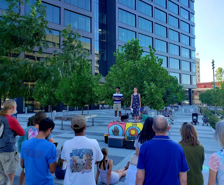 Picture of two high school students standing on an outdoor table with banners on it speaking to a crowd of people listening