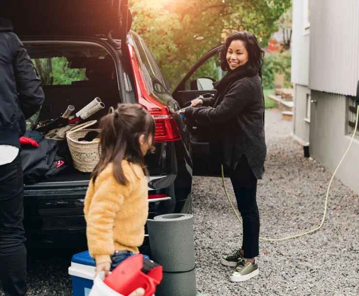 A family charges their black car on the side of a building with the trunk open after camping