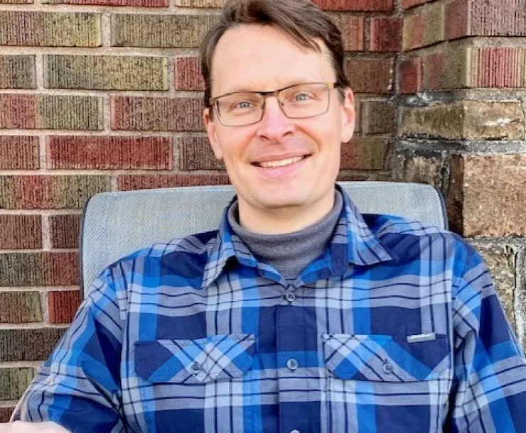 A man with short brown hair and glasses sat outdoors with a brick wall backdrop. He is smiling and wearing a blue plaid shirt.