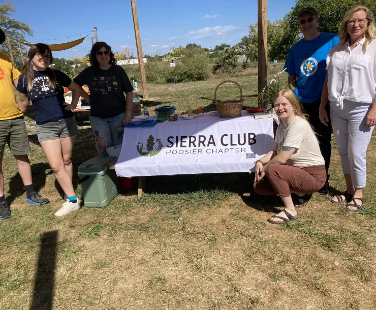 A group of six people outdoors on a blue-sky day, standing around a picnic table with a white banner with the Sierra Club Hoosier Chapter logo on it. They are all smiling and looking at the camera.