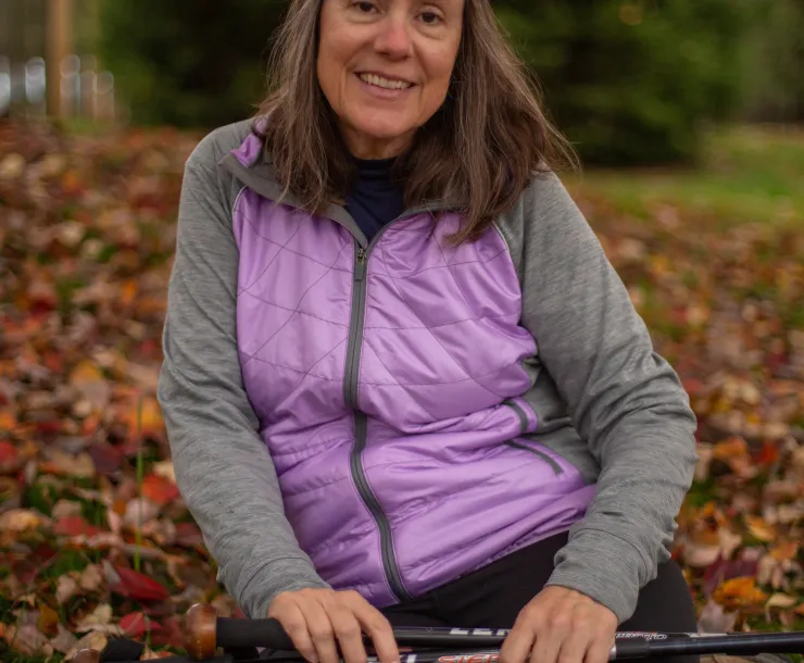 A woman with brown hair siting outdoors with a backdrop of fall leaves. She is wearing a purple windbreaker and a gray long sleeved shirt. She is smiling.