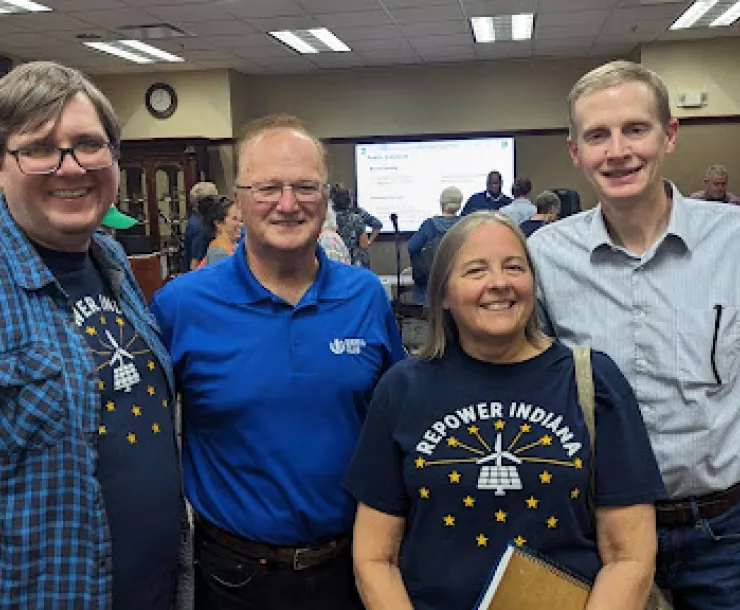 Four people stand at an indoor venue, smiling for the camera. Two of them are wearing blue t-shirts that state Repower Indiana.