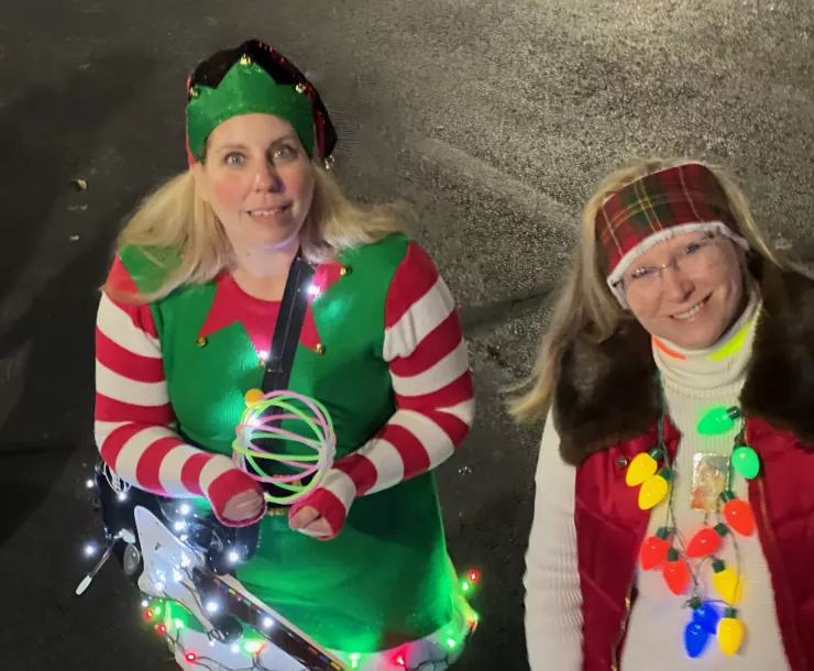 Two women with blonde hair wearing Christmas attire. One has a green elf hat and a green and red elf outfit. The other has string Christmas lights around her neck.