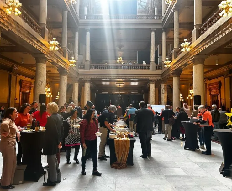 A large hall-type room with pillars and beams of light shining through. There are several people standing around, talking, and at tables.