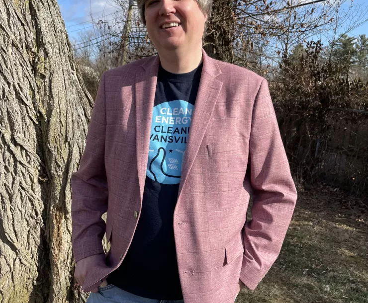 A person stood wearing a pink jacket and blue pants. They are outdoors with grass and some trees behind them. They are smiling and look relaxed.