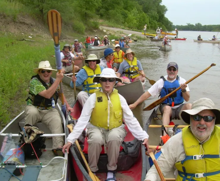 men and women wearing life jackets in canoes at river bank