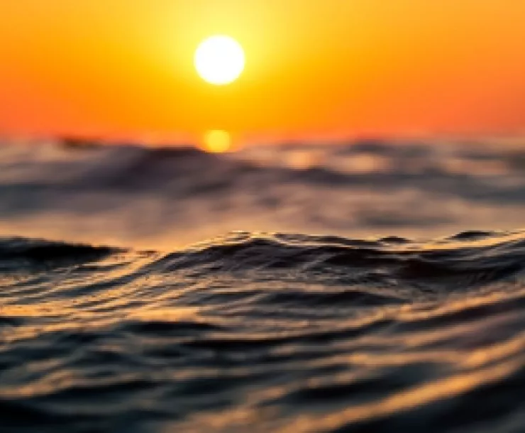 A bright yellow sun sets against a bright orange sky as ocean waves form in the foreground.