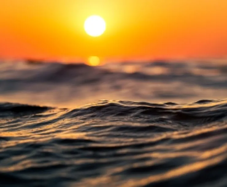 A bright yellow sun sets against a bright orange sky as ocean waves form in the foreground.
