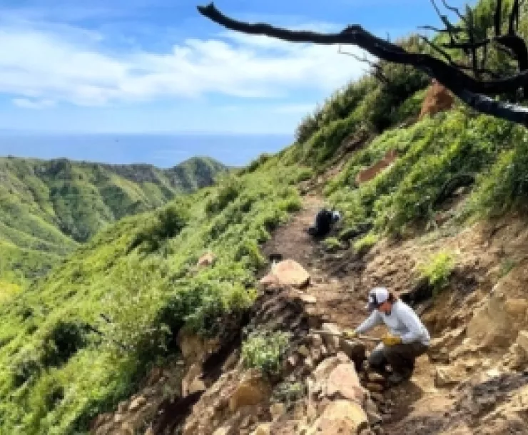 Woman kneels to inspect mountain trail after weather damage.