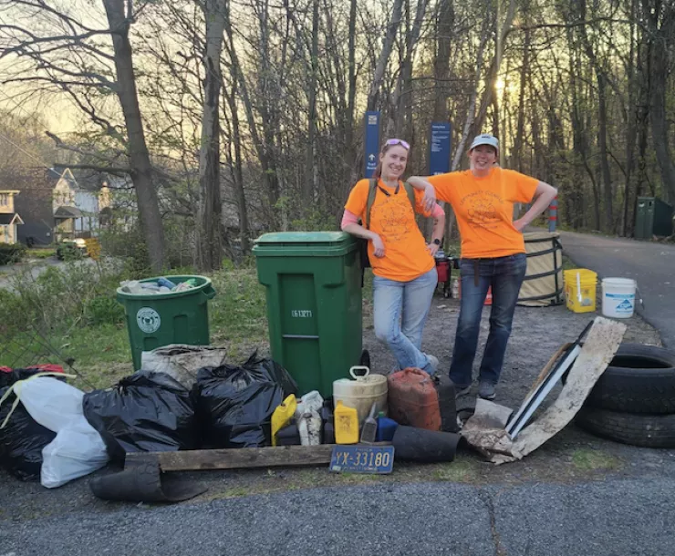 Two people pose next to trash bags and tires at a trailhead with houses in the back left.