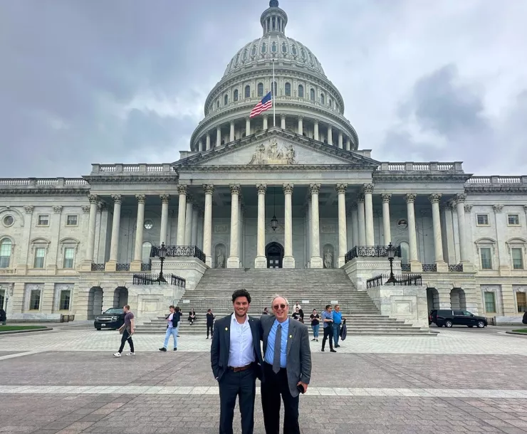 Two men stand in front of the Capitol Building in D.C. The building has a domed roof and several pillars at the front. The sky is cloudy.