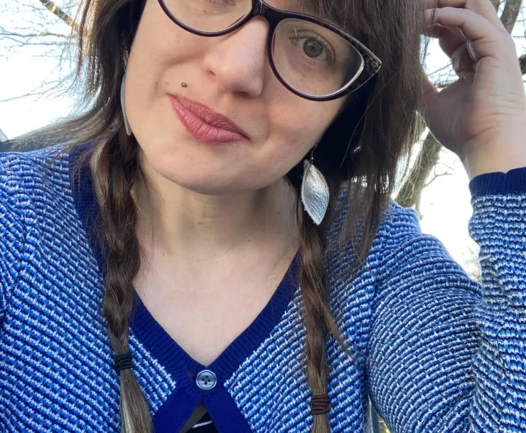 A woman with long brown hair in two plaits. She is wearing dark-rimmed glasses and a blue and white cardigan. There is a blue sky and a bare tree just visible behind her. She is smiling.