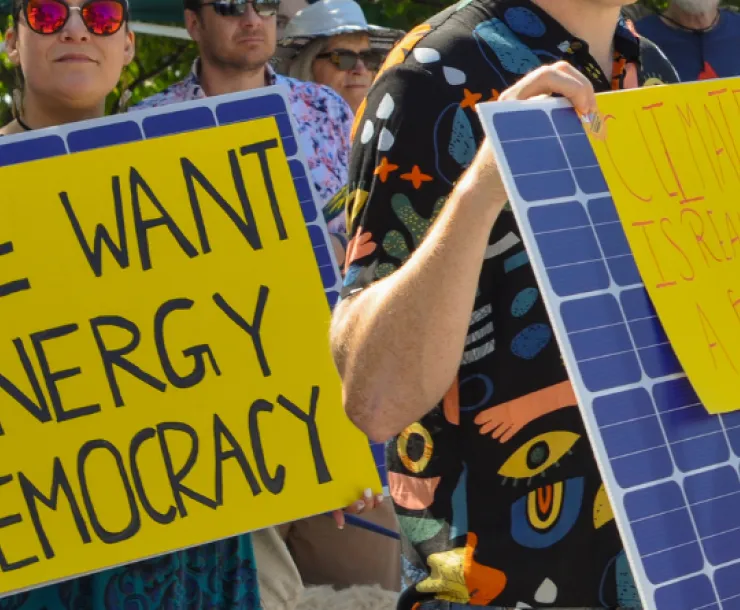 woman outdoors holding yellow sign that has the words "we want energy democracy" written in black. there are people behind her and a man to her right with a sign designed to look like a solar panel.