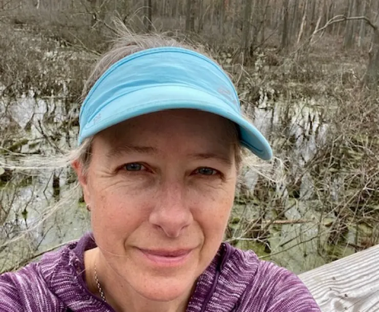 A head and shoulders photo of a person with a blue sports cap and a purple sweater, outdoors in the winter. The trees are bare. The person is smiling.