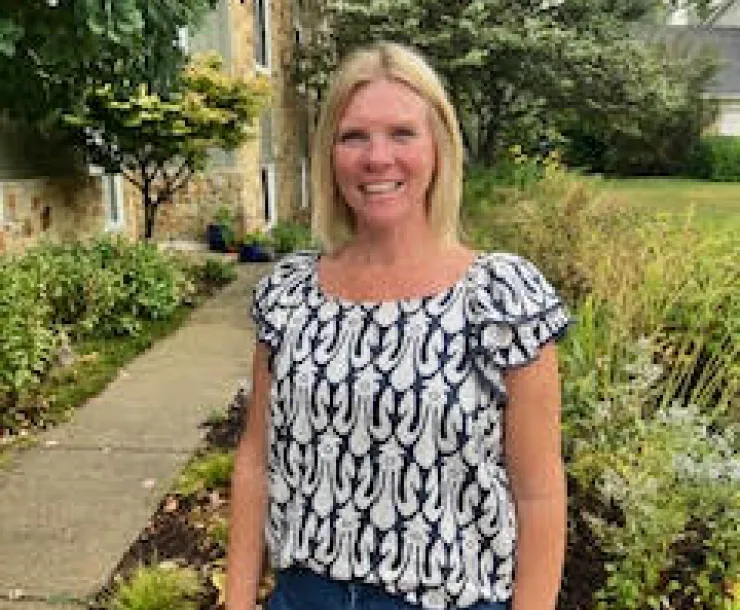 A woman with blonde hair stood in a yard with green plants and green grass behind her. She's wearing blue jeans and a black and white top and is smiling